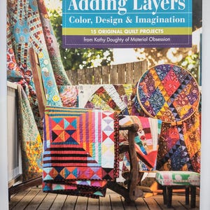 Puede incluir: Portada del libro "Adding Layers" con el subtítulo "Color, Design & Imagination". La imagen muestra coloridas colchas y telas. El libro promete 15 proyectos de colchas originales.
