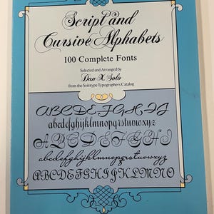 May include: A softcover book titled Script and Cursive Alphabets: 100 Complete Fonts, selected and arranged by Dan X. Solo. The cover is light blue with a white rectangular center featuring black text and various cursive alphabet samples.