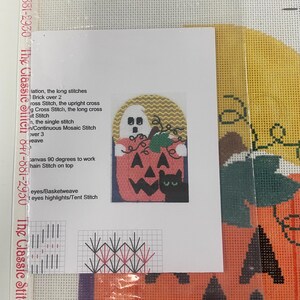 May include: A needlepoint canvas featuring a Halloween design with a large orange pumpkin, a white ghost, and a black cat. The canvas includes a printed instruction sheet with stitch diagrams and text, plus a barcode label at the top edge.