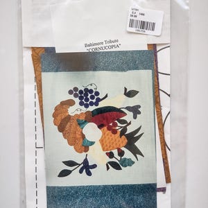 May include: A clear plastic bag containing a fabric panel with a colorful floral design, labeled "Baltimore Tribute 'CORNUCOPIA'". The design features a cornucopia overflowing with fruit and flowers. A white tag with a barcode is also visible.