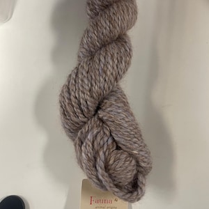 May include: A skein of Fauna yarn in a muted brown color with subtle blue and white flecks. The yarn is thick and textured, with a tag that reads "Fauna animal origins" and "The Fibre Company". Ideal for knitting and crochet projects.