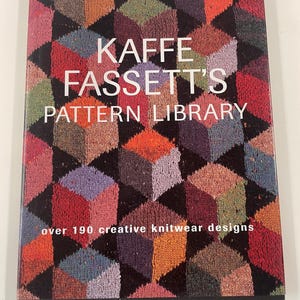 May include: A hardback book titled Kaffe Fassett's Pattern Library, over 190 creative knitwear designs. The cover features a repeating geometric cube pattern in shades of red, orange, purple, blue, and green, with white text centred on the front.