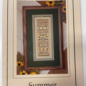 May include: A framed needlework design titled "Summer" by The Drawn Thread. The design features a cross-stitch pattern with floral motifs and the text "In a summer garden when days are sweet and slow." The frame is brown with a green mat.