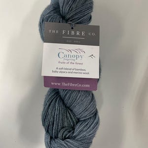 May include: A skein of blue-grey yarn with a tag from The Fibre Co. The tag reads "Canopy fingering fruits of the forest" and lists the yarn as a soft blend of bamboo, baby alpaca, and merino wool. The website www.TheFibreCo.com is also listed.
