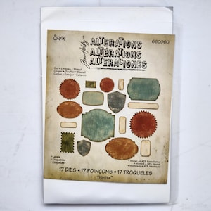 May include: A Sizzix Tim Holtz Alterations Thinlits die set package containing 17 metal dies for cutting, embossing, and stenciling. The packaging displays various label shapes in shades of red, green, blue, and tan, with the text 17 DIES.
