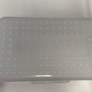 Puede incluir: Un estuche rectangular translúcido con una textura punteada en la tapa. La palabra "SpaceMaker" está grabada en la parte superior. El estuche está hecho de plástico transparente de color claro.