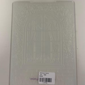 Peut inclure: Un dossier de gaufrage rectangulaire en plastique translucide présentant un motif en relief d'une fenêtre arquée avec des rideaux, entouré de motifs floraux et de vignes complexes. Une étiquette blanche avec un code-barres est collée.