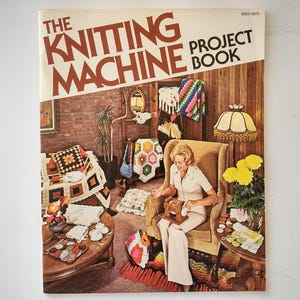 May include: A softcover book titled The Knitting Machine Project Book. The cover features a photograph of a person sitting in a tan armchair using a small knitting machine, surrounded by various crocheted and knitted items in a room setting.