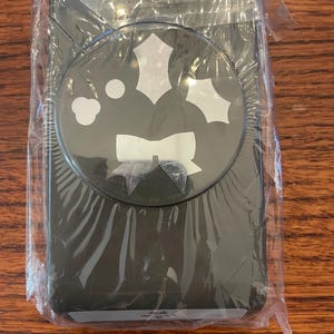 May include: A black paper punch tool encased in clear plastic packaging. The top of the device features a circular graphic displaying white silhouettes of a bow, holly leaves, and small circular shapes, indicating the punch-out designs it creates.