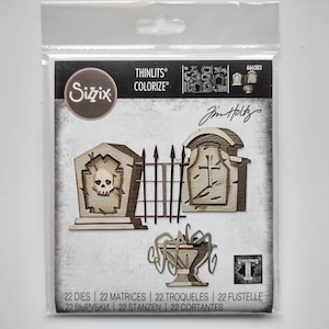 May include: A Sizzix Thinlits Colorize die set in a clear plastic package featuring 22 metal cutting dies. The packaging displays illustrations of two tombstones, a metal fence, and a decorative urn, with the text Sizzix, Thinlits, Colorize, and Tim Holtz.
