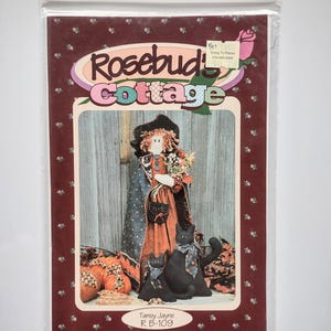 Può includere: Un pacchetto di cartamodelli Rosebud Cottage, modello R B-109 di Tansy Jayne, con una bambola e due gatti neri. La copertina mostra una bambola con un mantello scuro che tiene dei fiori, con due gatti neri seduti accanto. Il testo include Rosebud Cottage, Tansy Jayne, R B-109 e i dettagli dell'indirizzo.