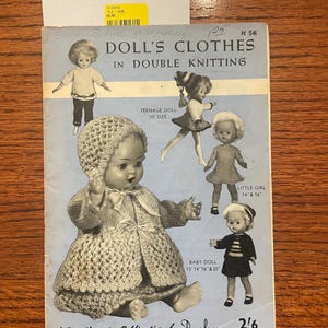 May include: A vintage instructional booklet titled DOLL'S CLOTHES IN DOUBLE KNITTING, N 56, A Needlecraft Publication by Penelope. The light blue cover features black and white illustrations of dolls wearing various knitted outfits with size labels.