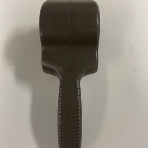 May include: A dark brown plastic handheld tool featuring a wide, rounded top section and a narrower, textured handle with horizontal ridges. The object has a smooth, molded finish with a visible seam running vertically down the center of the handle.