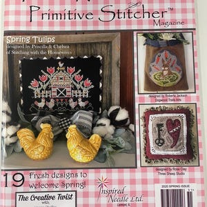 以下が含まれることがあります： ピンクと白のギンガムチェックの背景を持つ「Punch Needle & Primitive Stitcher」の雑誌の表紙。表紙には、納屋のシーン、ニンジンを持つウサギ、鍵とハートなど、春をテーマにしたニードルワークのデザインが掲載されています。雑誌は2020年春号です。
