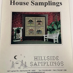 May include: A printed paper with the title "House Samplings" featuring a cross-stitch design of four houses. The design is framed and set against a brick wall background. The paper also includes the text "HILLSIDE SAMPLINGS".