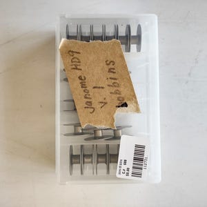 Könnte beinhalten: Ein rechteckiger, durchscheinender Kunststoff-Aufbewahrungsbehälter mit mehreren silberfarbenen Nähmaschinenspulen aus Metall. Auf der Oberseite ist ein zerrissenes Stück beiges Papier mit dem handschriftlichen Text Janome HP9 v.1 bobbins aufgeklebt.