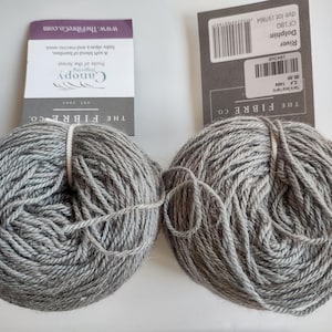 May include: Two grey yarn skeins with labels from The Fibre Co. The labels show the yarn name "Dolphin" and "Canopy". The yarn is a blend of bamboo, baby alpaca, and merino wool. The skeins are round and have a soft, textured appearance.
