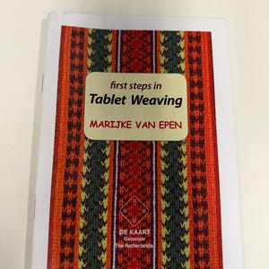 May include: A softcover instructional book titled first steps in Tablet Weaving by Marijke van Epen. The cover features a vibrant, vertical striped pattern in shades of red, orange, yellow, and green, with a small logo for De Kaart Gelselaar.
