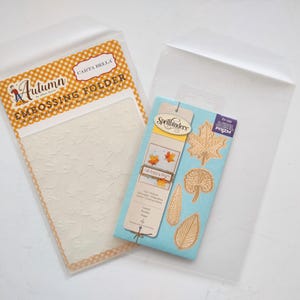 May include: A Carta Bella Autumn embossing folder with a leaf pattern and a Spellbinders die set featuring four distinct leaf shapes on a light blue backing card, both items enclosed in clear plastic protective sleeves for crafting projects.