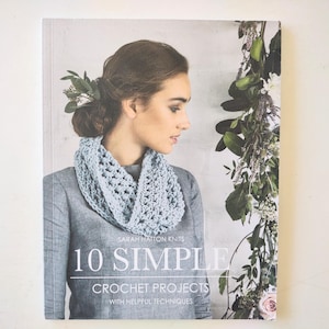 May include: A softcover book titled 10 Simple Crochet Projects with Helpful Techniques by Sarah Hatton Knits. The cover features a person wearing a light blue crocheted cowl over a grey shirt, with green foliage visible on the right side.
