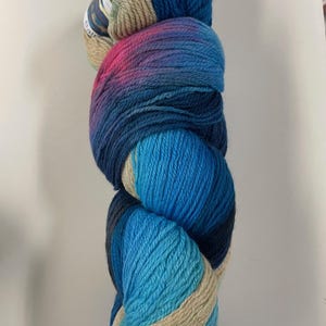 May include: A skein of colorful yarn with a gradient of blues, pink, and tan. The yarn is tightly wound, showing the texture of the fibers. A label is attached at the top. Ideal for knitting or crocheting projects.