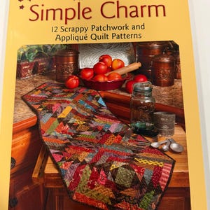 May include: A softcover book titled Simple Charm by Kim Diehl, featuring 12 Scrappy Patchwork and Appliqué Quilt Patterns. The cover displays a colorful, multi-patterned fabric table runner on a wooden surface with copper canisters and fruit.