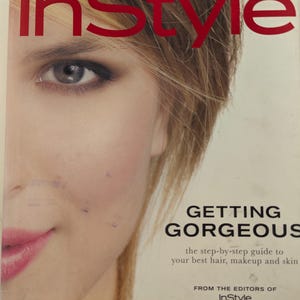May include: A softcover book titled InStyle Getting Gorgeous, the step-by-step guide to your best hair, makeup and skin from the editors of InStyle. The cover features a partial close-up of a face with blonde hair, blue eyes, and pink lips.