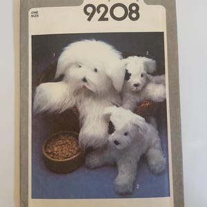 Pode incluir: Um envelope de molde de costura Simplicity 9208 com ilustrações de três cães de pelúcia. Um cão branco grande e felpudo está sentado numa cesta, enquanto dois cães brancos menores com manchas pretas nas orelhas são mostrados.