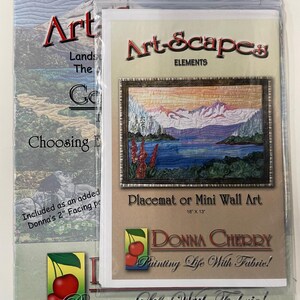May include: A package containing a placemat or mini wall art. The art features a landscape scene with mountains, water, and trees. The package includes the text "Art-Scapes Elements" and "Donna Cherry Painting Life With Fabric!" The placemat measures 18" x 13".