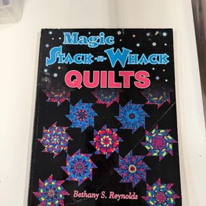 May include: A softcover instructional book titled Magic Stack-n-Whack QUILTS by Bethany S. Reynolds. The black cover features a repeating pattern of colorful, kaleidoscopic star-shaped quilt blocks in shades of blue, pink, and yellow.