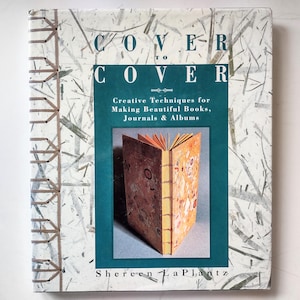 May include: A hardcover book titled Cover to Cover: Creative Techniques for Making Beautiful Books, Journals & Albums by Shereen LaPlantz. The cover features a white textured paper with visible fibers and a visible brown thread spine binding.