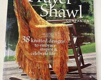 May include: A book titled "The Prayer Shawl Companion" with a shawl draped over a wooden rocking chair. The shawl is orange and yellow with fringe. The book cover has text that reads "38 knitted designs to embrace inspire & celebrate life."