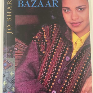 Könnte beinhalten: Buchcover von "Knitting Bazaar" von Jo Sharp. Es zeigt eine Frau in einem lila Strickcardigan mit einem Muster in Schwarz, Rot und Gold. Sie trägt ein gelbes Hemd mit Kragen. Der Buchtitel ist blau.