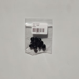 Puede incluir: Una pequeña bolsa de plástico transparente que contiene una colección de pequeñas cuentas negras redondas. Una etiqueta blanca con texto y un código de barras está adherida a la bolsa. La etiqueta dice "Vintage Jet Glass Buttons" y "C.# 1375".