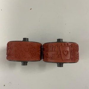 May include: Two cylindrical rubber stamps with a dark reddish-brown finish. The surface of the stamps features embossed designs including coffee cups, hearts, cupcakes, and small floral patterns, each mounted on a central metal axle for rotation.