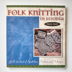 Może przedstawiać: Książka w miękkiej oprawie zatytułowana Folk Knitting in Estonia autorstwa Nancy Bush. Okładka zawiera historyczne zdjęcie dziecka w odcieniach niebieskiego oraz prostokątną wstawkę z trzema ręcznie robionymi skarpetami w czarno-białe wzory.