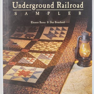 May include: A book titled "Underground Railroad Sampler" by Eleanor Burns & Sue Bouchard. The cover features a quilt with various geometric patterns in shades of brown, red, and blue. A lit lantern sits on a map in the background.