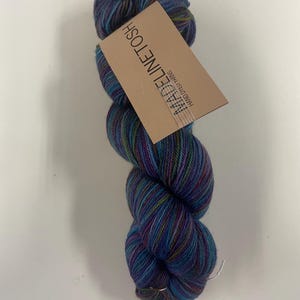 May include: A skein of hand-dyed yarn featuring a variegated color palette of deep blue, purple, and hints of green. A rectangular tan paper label is attached to the yarn, printed with the text MADELINETOSH and the words HAND DYED YARNS.