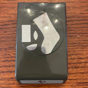 May include: A dark grey paper punch tool featuring a circular top section with silver-coloured cutouts of a sock, a small rectangle, and two curved shapes. The base of the tool has a rectangular grey label with a small icon of a sock printed on it.