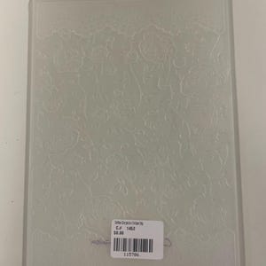 Puede incluir: Una carpeta de relieve rectangular de plástico translúcido con un patrón floral intrincado en relieve con rosas y un borde similar al encaje en la parte superior. Una etiqueta rectangular blanca con un código de barras y el texto C.# 1452 está adherida en el centro.