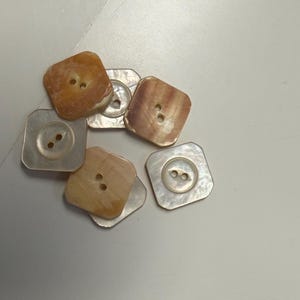 May include: A collection of six square-shaped buttons with rounded corners, featuring a pearlescent finish in shades of tan, beige, and cream. Each button has two central holes for attachment and a smooth, slightly iridescent surface texture.