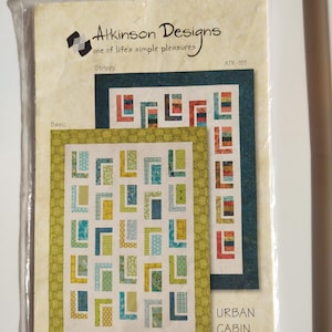 May include: A quilting pattern package by Atkinson Designs, featuring the "Urban Cabin" design. The pattern showcases two quilt designs with geometric patterns in blue, green, yellow, and orange. The text "Strippy" and "Basic" are also visible on the package.