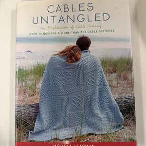 以下が含まれることがあります： メリッサ・リープマン著のソフトカバー本『Cables Untangled: An Exploration of Cable Knitting』。表紙には、様々なケーブル編みの模様が施された水色のニットブランケットが、ビーチに座る二人の上に掛けられている様子が写っています。