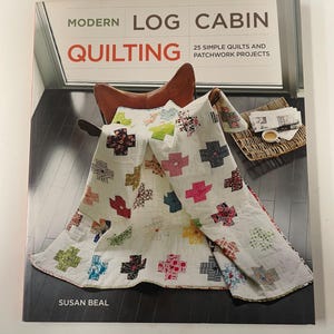 May include: A book titled Modern Log Cabin Quilting: 25 Simple Quilts and Patchwork Projects by Susan Beal. The cover features a white quilt with colourful patterned cross-shaped blocks draped over a brown leather chair on a dark wood floor.