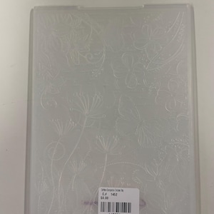 Puede incluir: Una carpeta de relieve de plástico transparente con un patrón floral y de mariposas en relieve. La superficie muestra delicados dibujos de líneas de flores y una mariposa. Una etiqueta rectangular blanca está pegada en la parte inferior con texto y un código de barras.