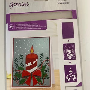 May include: A 3-piece Gemini embossing folder and stencil set. The package features a festive candle design with a red candle, a red bow, and green holly. The card measures 5" x 7" (12.7cm x 17.8cm).