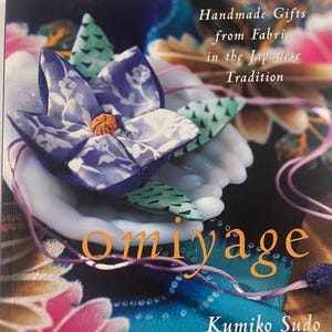 May include: A book cover with the title "Handmade Gifts from Fabric in the Japanese Tradition." The cover features a fabric flower in shades of blue and white, with the word "Omiyage" in orange. The author's name, Kumiko Sudo, is also visible.