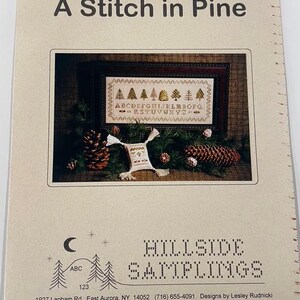 May include: A printed pattern titled "A Stitch in Pine" featuring a framed cross-stitch design with pine trees and an alphabet. The image includes pine cones and greenery, with the text "HILLSIDE SAMPLINGS" and contact information.