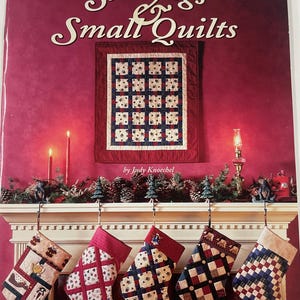 May include: A book cover titled "Stockings & Small Quilts" by Judy Knoechel. The cover features a small quilt and a fireplace mantel decorated for Christmas with stockings hanging from it. The stockings have various patterns and colors.