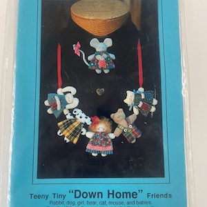 May include: A blue package featuring a black background with several small fabric dolls. The dolls include a mouse, bear, rabbit, and girl, each wearing a dress. The text reads "Teeny Tiny 'Down Home' Friends" and "Kitty Lou's Creations". The dolls are approximately 2.5 inches tall.